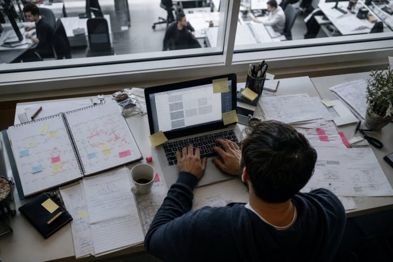 A messy desk holds a laptop, coffee cup, calendar, and scattered printed bug reports.
