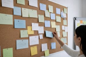 A cork noticeboard covered in colorful sticky notes and cards, with a hand pinning a fresh one.