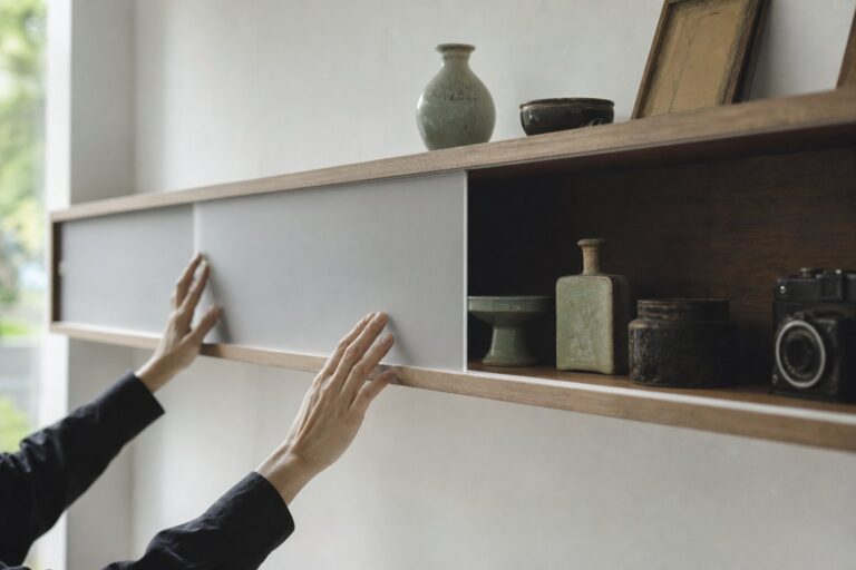 Hands slide a frosted glass panel across a wooden shelf, partly hiding a row of vintage objects from view.