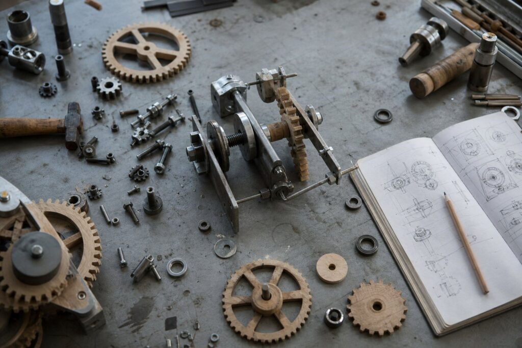 A workbench seen from above holds steel bolts, wooden gears, a half-built device, and an open sketchbook with a pencil.