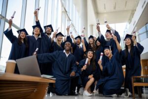 Graduates in caps and gowns laugh and raise rolled certificates in a sunlit university common area.
