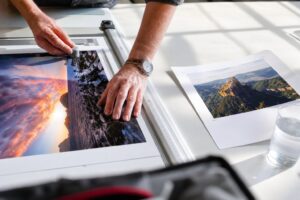 Hands trim a photo with a rotary cutter on a white mat beside another print with pencil marks.