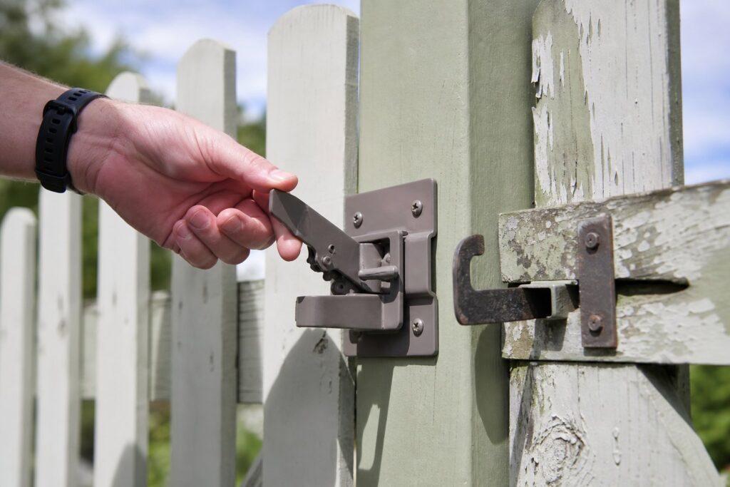 A hand tests a new metal latch on a freshly painted wooden fence post outdoors.