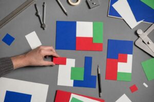 A hand arranges colorful geometric paper cutouts on a grey workbench beside drafting tools and a stylus, seen from above.