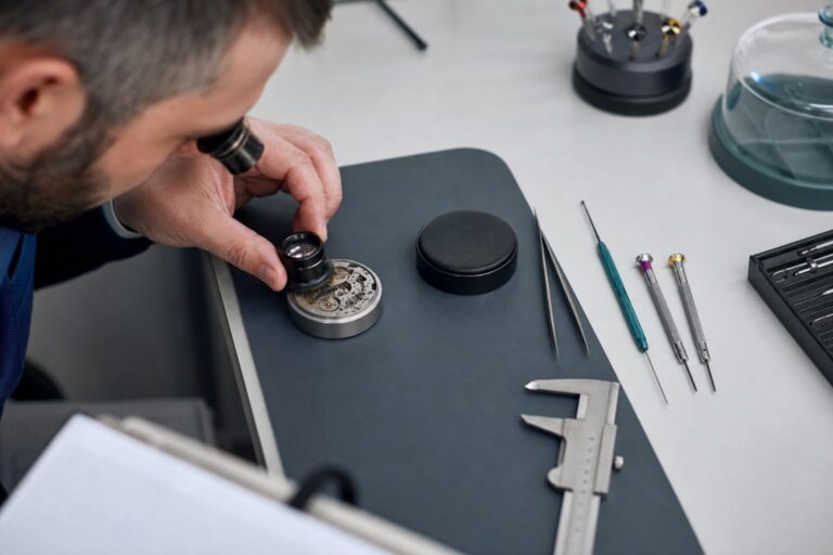 A technician's hand holds a loupe over a disassembled mechanical part on a tidy workbench lined with precision tools.