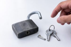 A heavy steel padlock sits unlatched on a white surface next to small keys, one bent, as a hand reaches toward it.