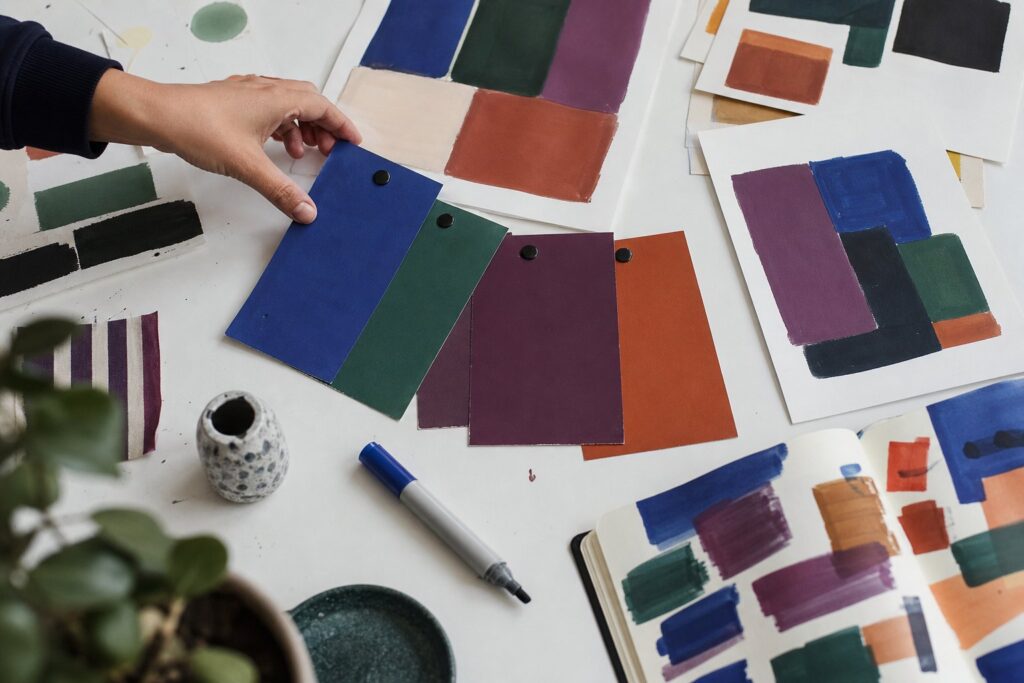A hand reaches toward colorful jewel-toned swatches fanned on a white table beside sketches, a marker, and an open notebook.