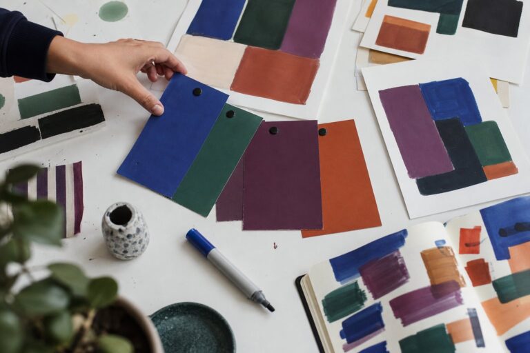 A hand reaches toward colorful jewel-toned swatches fanned on a white table beside sketches, a marker, and an open notebook.