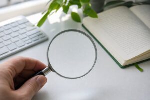A hand holds a magnifying glass over a desk, sharpening text into focus beside a keyboard and open notebook.